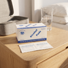 Alcohol Wipes 100 Pack box and two 70% isopropyl alcohol pad sachets on a wooden bedside table next to a glass of water and a grey toiletry case