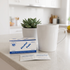 Alcohol Wipes 100 Pack box and two 70% isopropyl alcohol pad sachets on a light stone kitchen bench beside a white mug and a potted succulent